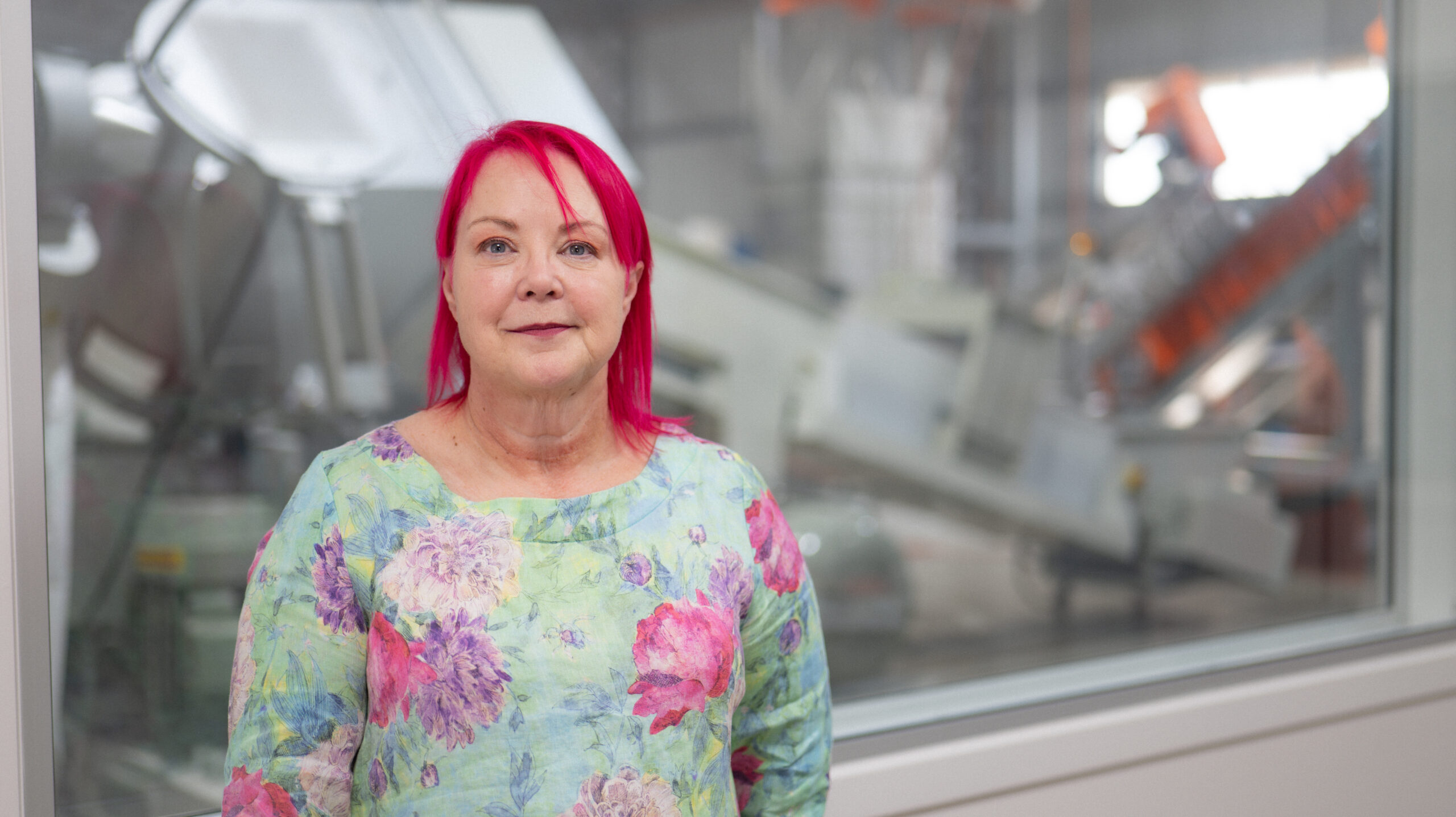 A woman with bright pink hair and a floral shirt is smiling and facing the camera. She is standing in front of machinery.